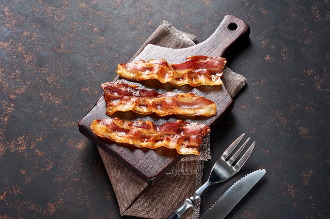 Trei bucati de bacon prajit asezate pe un tocator de bucatarie