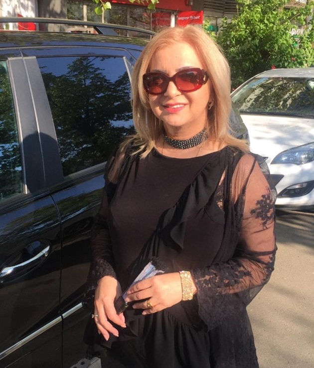 Carmen Șerban lângă o mașină neagră, îmbrăcată elegant, cu ochelari de soare pe ochi și ținând în mână un evantai