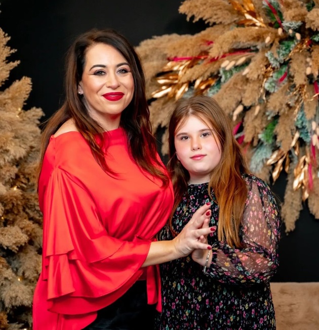 Oana Roman purtând o bluză roșie și fiica ei Isabela cu o cămașă în motive florale, ținându-se de mână în fața unui decor modern de crăciun 
