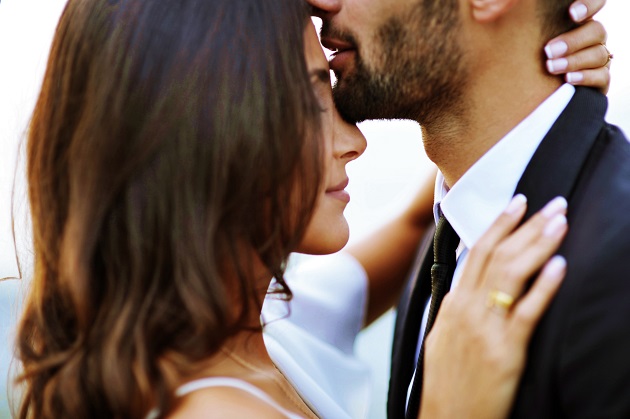 Un bărbat își sărută pe frunte iubita în timp ce aceasta îl mângîie pe gât și are ochii închiși.