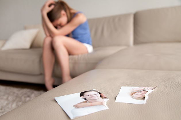 Fată tânără care stă și suferă singură pe o canapea în timp ce rupe pozele pe care le are împreună cu iubitul său. 