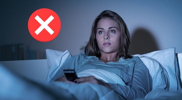 Femeie care se uită noaptea la televizor, în timp ce stă în pat cu telecomanda în mână