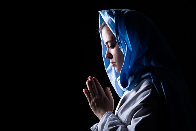Femeie care se roagă în întuneric, în timp ce poartă o eșarfă pe cap și ține mâinile împreunate