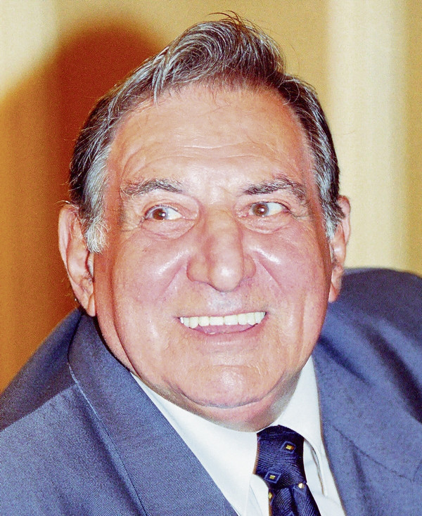 Jean Constantin îmbrăcat în costum accesorizat cu cravată, zâmbind și privind într-o parte