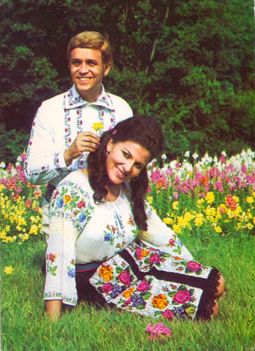 Benone Sinulescu și Irina Loghin în tinerețe, îmbrăcați în costume populare pe un câmp cu flori, în timp ce el îi punea o floare la ureche