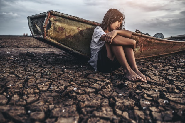 Fetiță afectată de secetă, care stă pe pământul uscat și crăpat, lângă o barcă