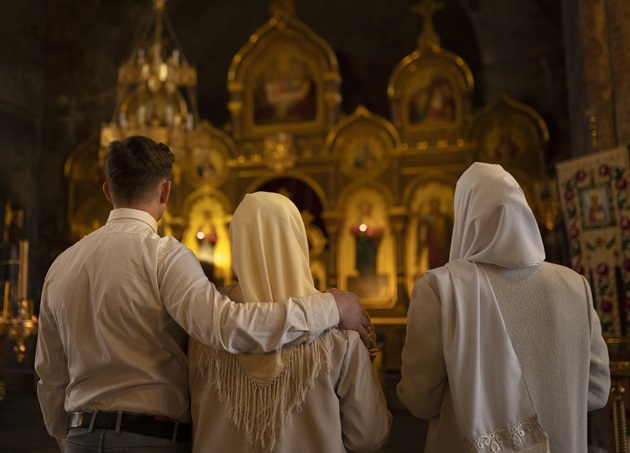 Două femei cu capul acoperit și un bărbat îmbrăcat în cămașă care se roagă într-o biserică