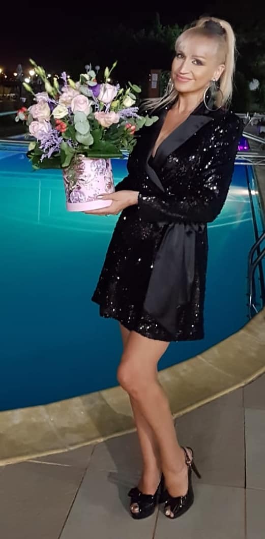 Simona Florescu într-o rochie neagră, scurtă, cu paiete, pe marginea piscinei, ținând în mână o cutie cu flori