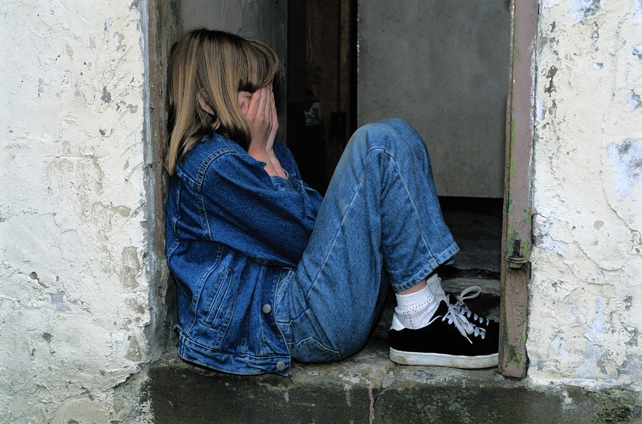 fetita blonda imbracata în jeansi care isi acopera fata in timp ce plange, asezata pe jos la intrarea intr-o casa in constructie