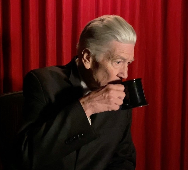 david lynch imbracat in negru pe fundal rosu bea dintr-o cana