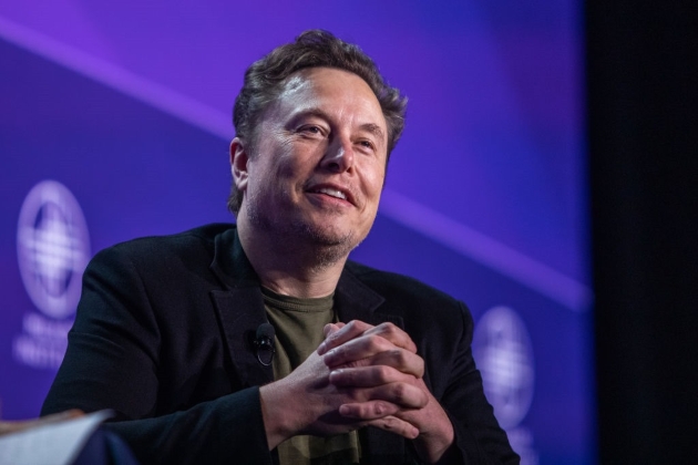 elon musk in cadrul unei conferinte de presa imbracat in negru, fundal albastru