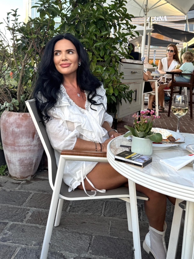 ioana nastase intr-o bluza alba si botine se afla la masa si serveste un pahar cu vin