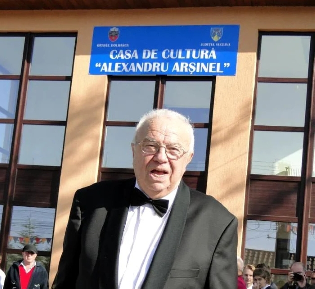 alexadru arsinel imbracat in costum negru si papion fotografie in fata casei de cultura din dolhasca care ii poarta numele