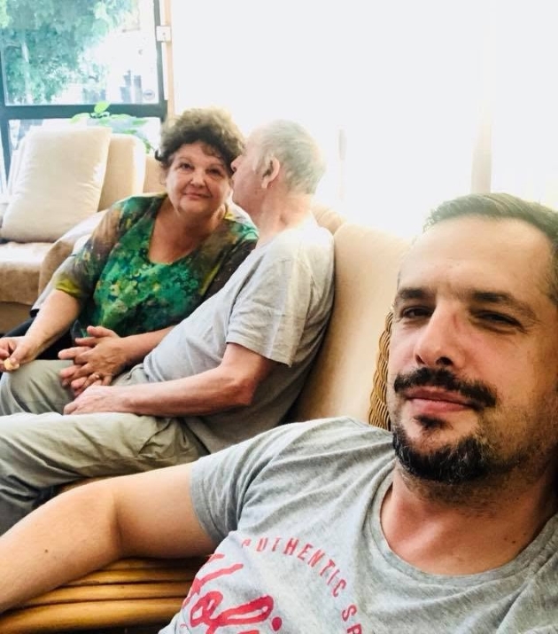 madalin ionescu alaturi de parintii sai in timp ce acestia stau pe canapea si tatal o saruta pe mama pe obraz