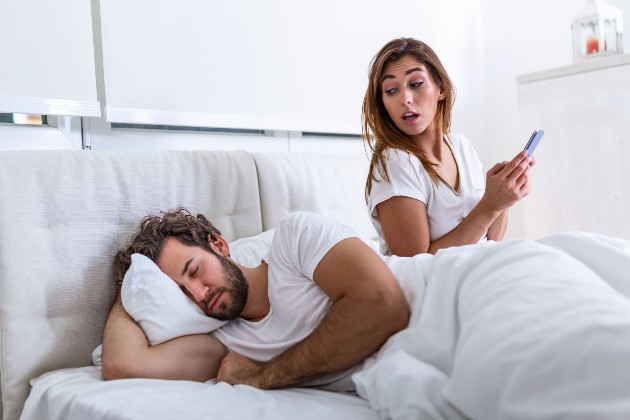 Imagine cu o femeie care verifică un telefon și cu un bărbat care doarme, amândoi în pat
