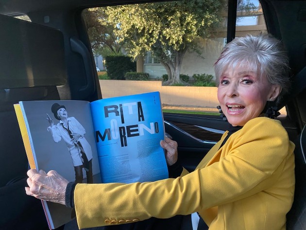 Imagine cu Rita Moreno într-un sacou galben, zâmbind în timp ce ține în mână o revistă cu numele ei