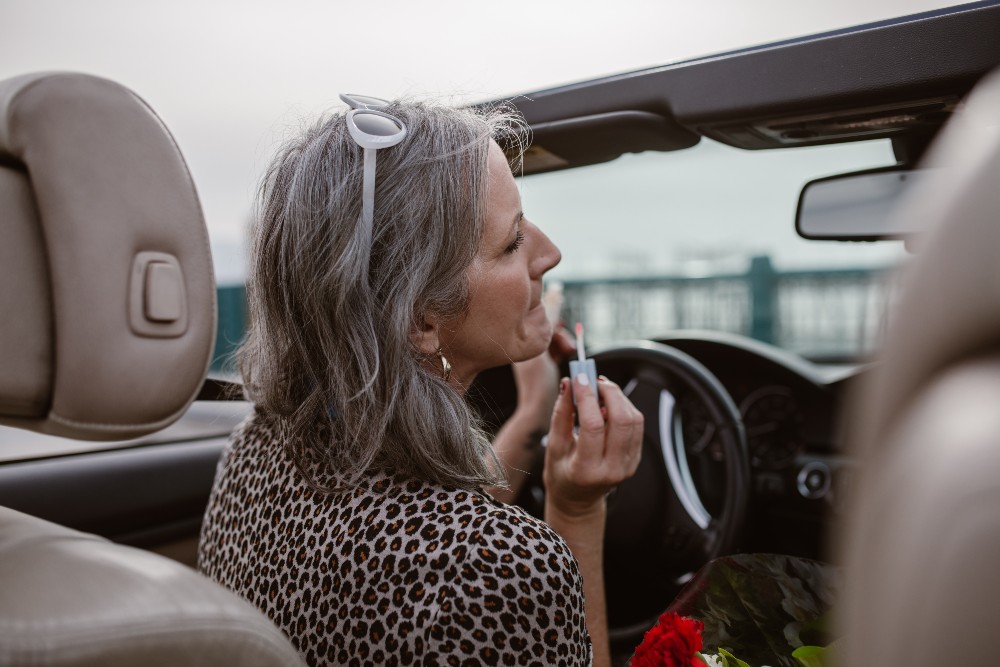 Imagine cu o femeie cu părul gri care se dă cu ruj în mașină