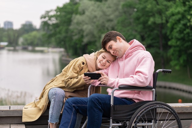 Imagine cu o femeie fericită și un bărbat într-un scaun cu rotile care se uită la un telefon