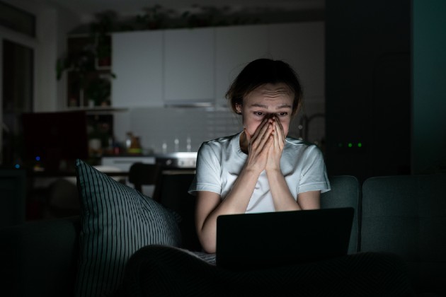 Imagine cu o femeie care stă pe canapea și se uită îngrozită către laptop