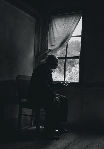Imagine alb-negru cu un bărbat bătrân care se uită pe geam
