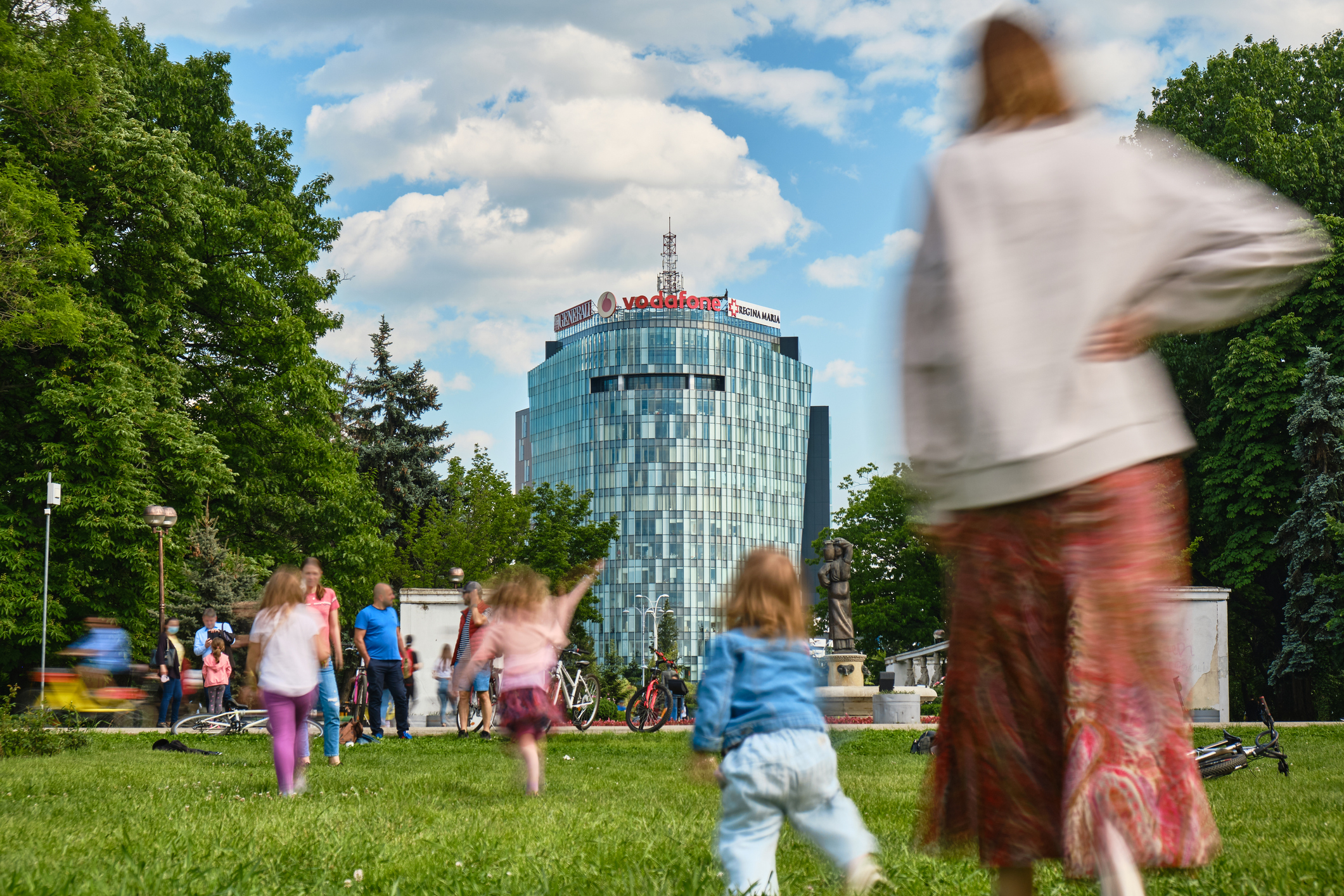 siluete de oameni in parc, iarba verde si copaci cu cerul insorit