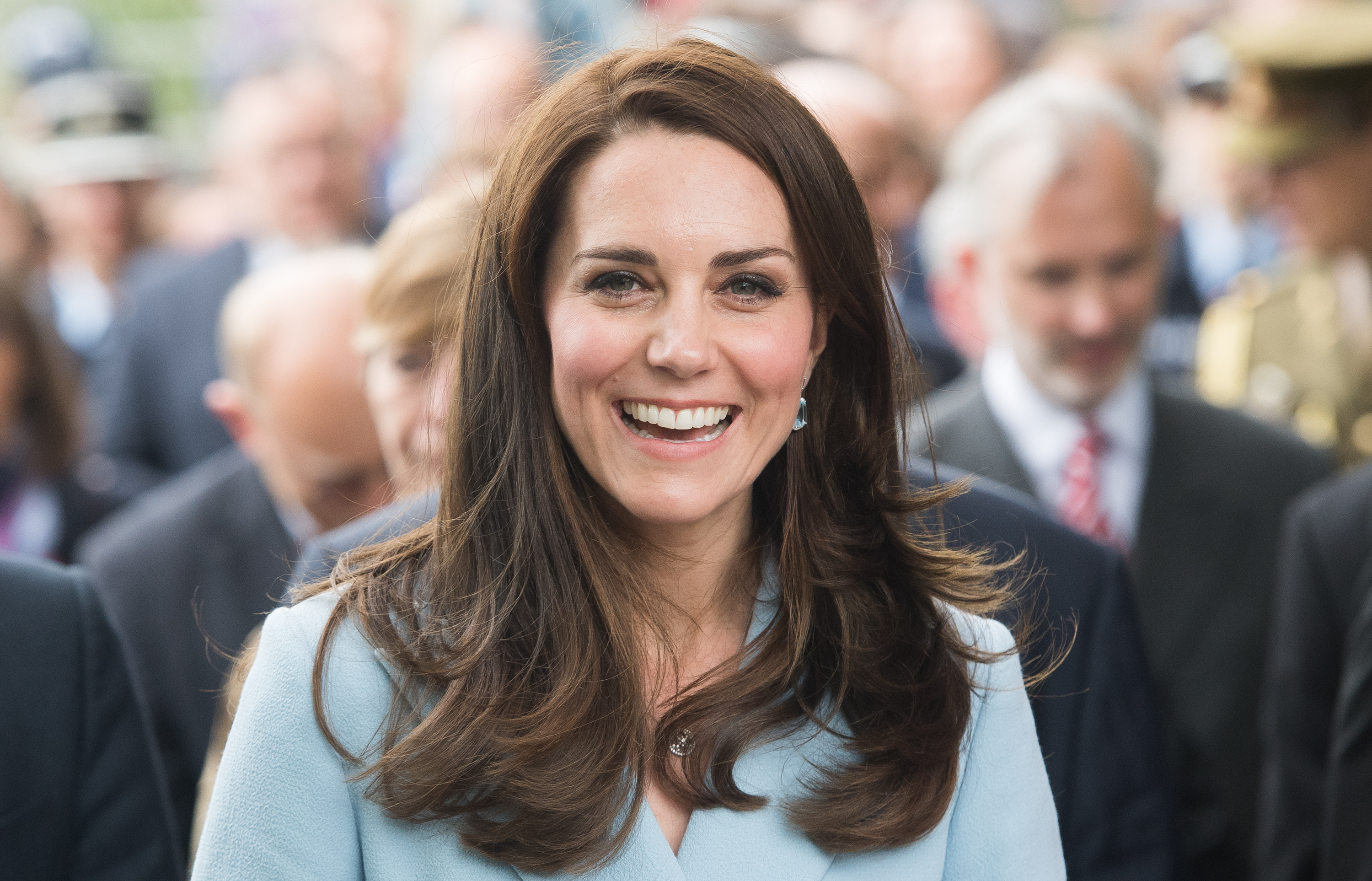 Kate Middleton fotografiata din fata, in timp ce zambeste larg. Poarta un trench bleu deschis 