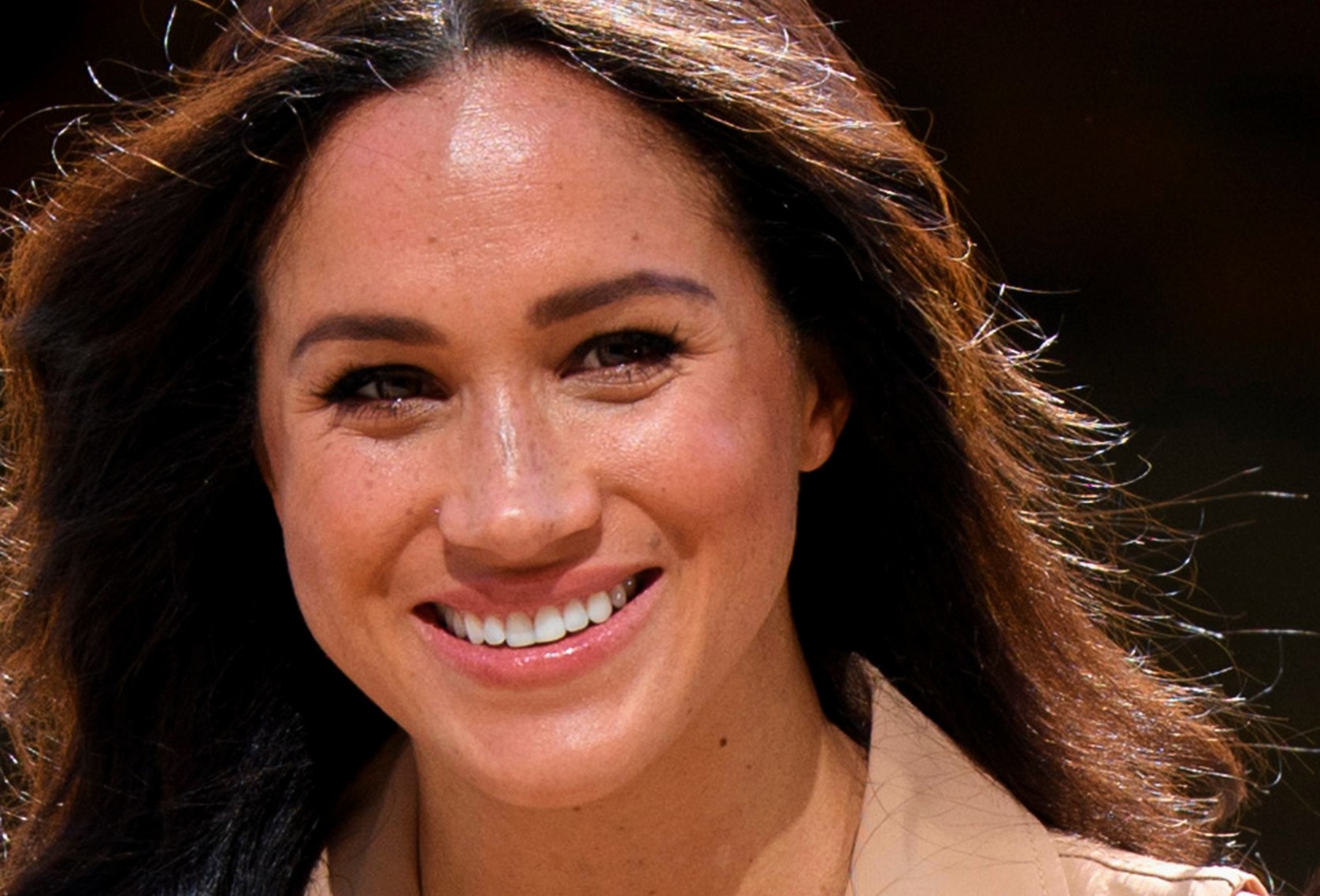 Meghan Markle zambeste larg in fata camerei, in timp ce priveste intr-o parte. Poarta un sacou bej, pe un fundal negru