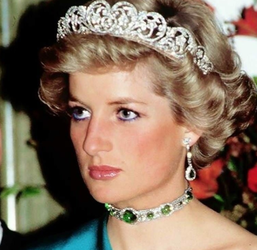 Printesa Diana se uita intr-o parte si zambeste subtil. Poartă o rochie albastră cu umarul dezgolit, o coroana cu diamante, dar și un colier și cercei cu pietre pretioase.