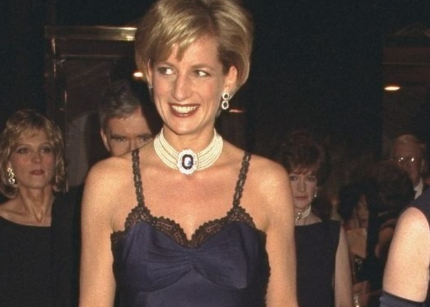 Printesa Diana priveste intr-o parte si zambeste larg. Poarta o rochie neagră cu bretele și accesorii cu perle si pietre pretioase