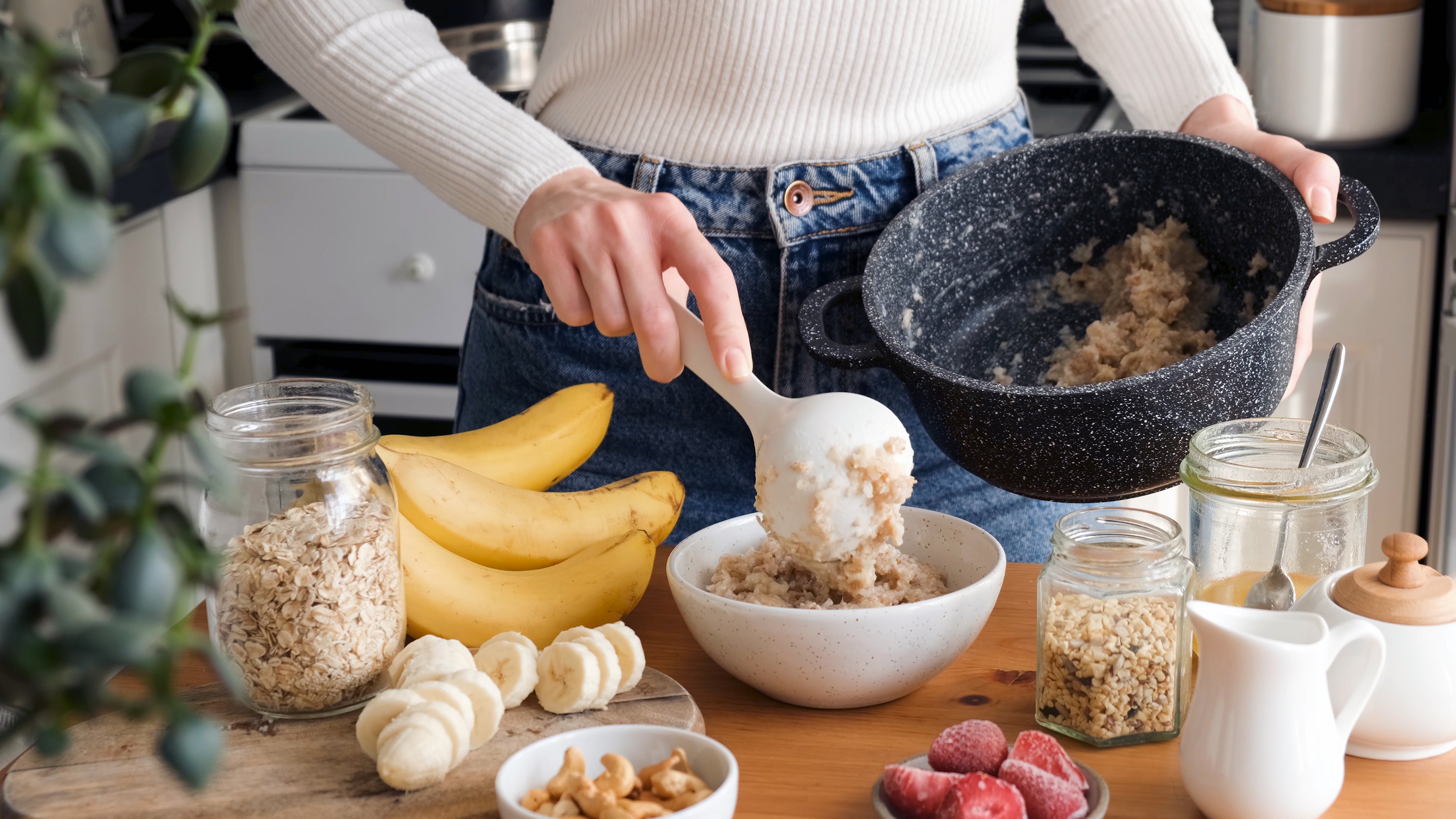 o femeie tine in mana o oala cu ovaz, iar cu cealalta pune terciul de ovaz intr-un bol alb, pe o masa de lemn, alaturi de un manunchi de banane