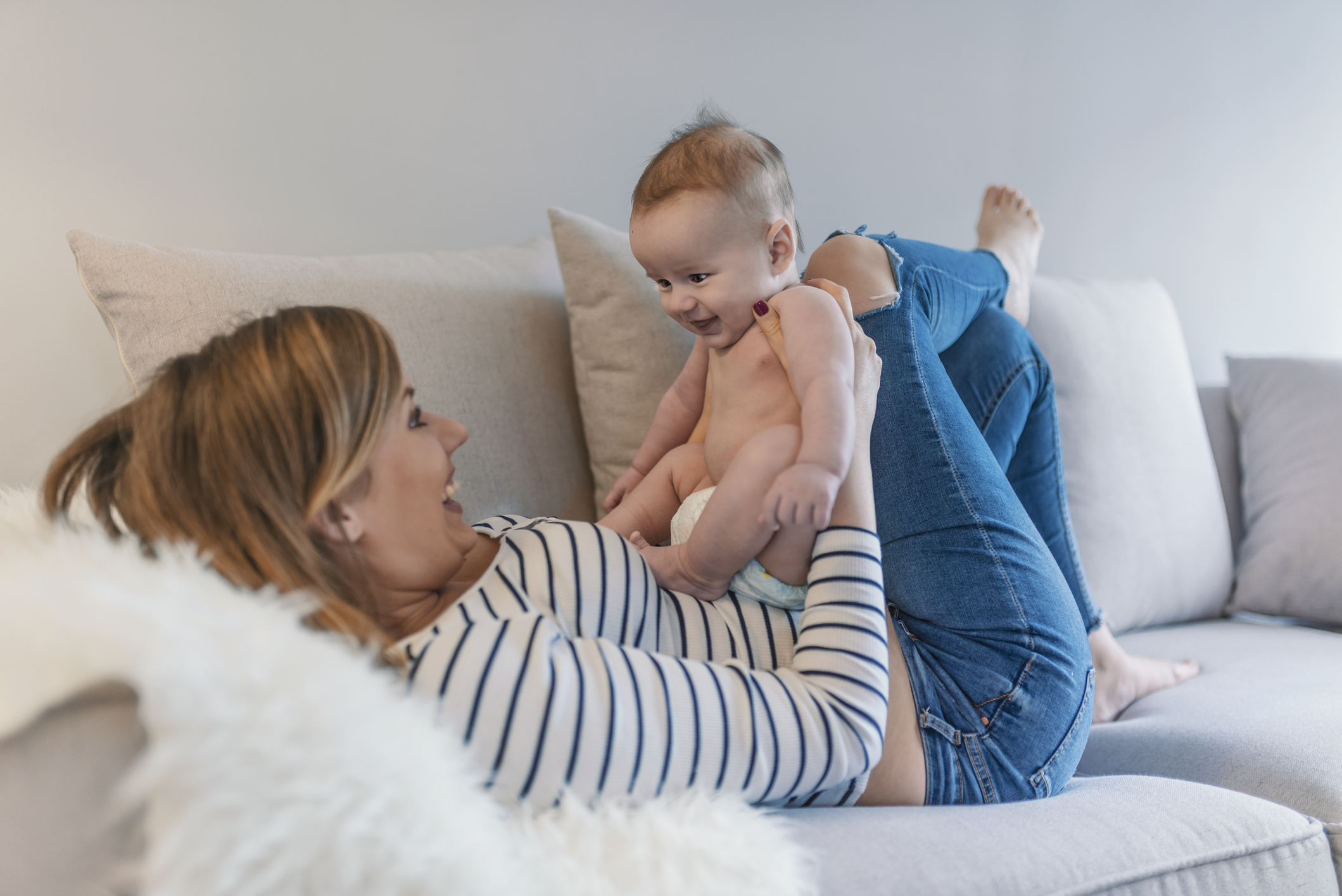 mama asezata pe spate pe canapea, in timp ce isi tine bebelusul pe burta, in sezut, ambii zambesc si se uita unul la celalalt