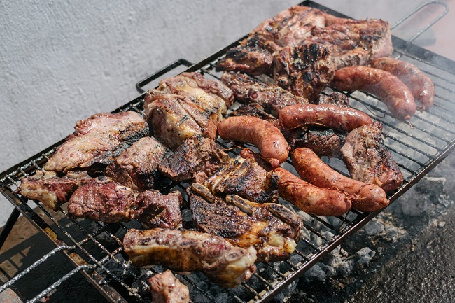 bucati de carne de porc si carnati asezate pe un gratar negru, cu carbuni incinsi din care iese fum