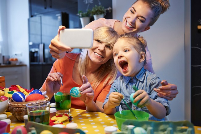 o mama, bunica si fiica fac o fotografie impreuna, in timp ce stau asezate la masa si picteaza oua de Paste