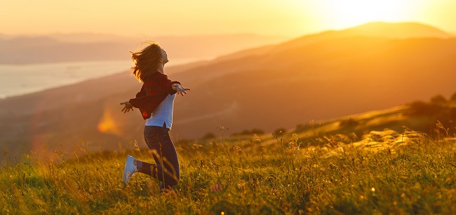 o femeie sare in sus de fericire, pe un camp inflorit, la apusul soarelui