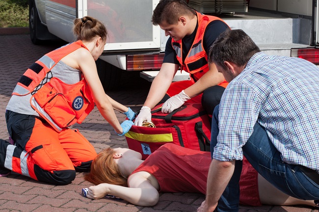 o femeie sta intinsa pe spate, alaturi de un barbat care sta langa ea si doi paramedici care scot instrumente medicale din trusa de prim ajutor