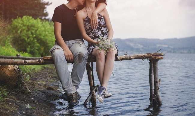 un cuplu sta pe o bucata de lemn, pe marginea unui lac, in timp ce el o tine in brate pe ea si incearca sa o sarute pe gat