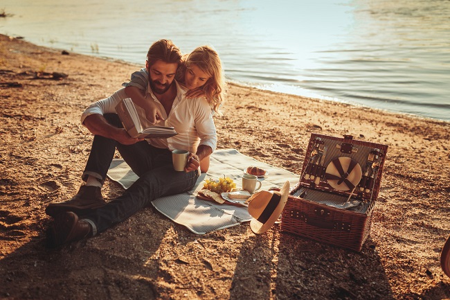  un barbat si o femeie stau imbratisati pe plaja, in timp ce citesc o carte si se bucura de un picnic