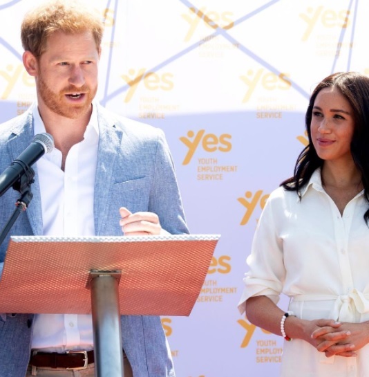 printul harry vorbeste in cadrul unei conferinte, in timp ce meghan markle il priveste dintr-o parte