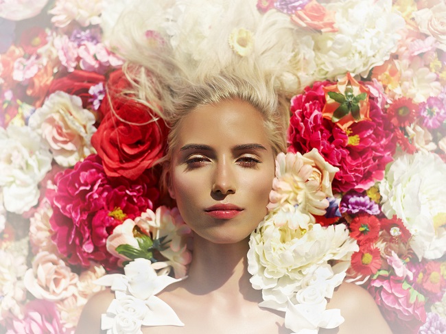  imagine de sus cu o femeie blonda, inconjurata de flori colorate care priveste fix in fata