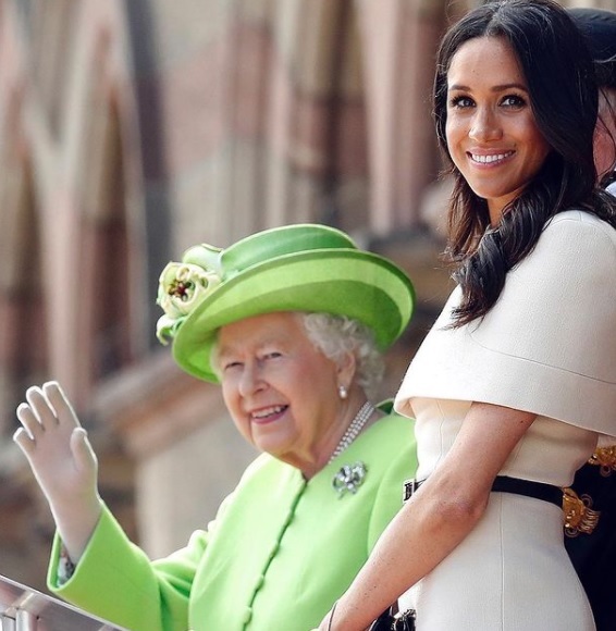 meghan markle priveste intr-o parte si zambeste, in timp ce regina elisabeta a II-a saluta multimea, in timpul unui eveniment