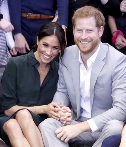 meghan markle si printul harry stau asezati pe scaun si zambesc larg