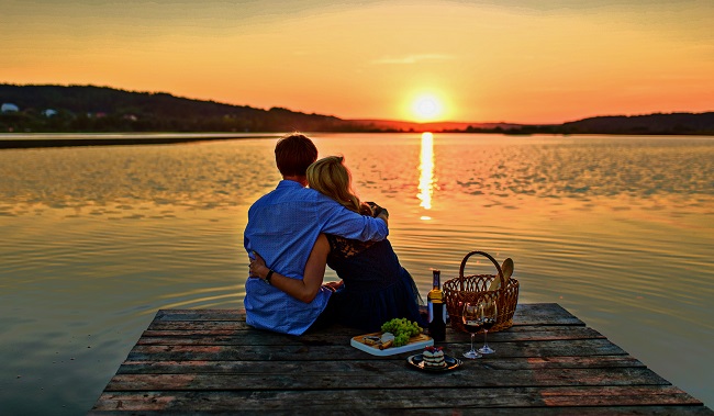 un barbat si o femeie stau imbratisati pe un ponton al unui lac si privesc apusul de soare