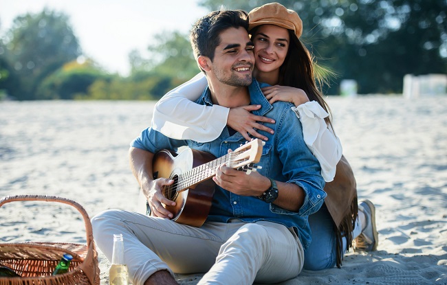 o femeie imbratiseaza un barbat si priveste intr-o parte, in timp ce el zambeste si canta la chitara