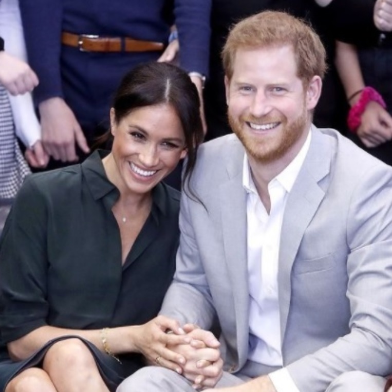 meghan markle si printul harry stau asezati pe scaun si zambesc larg