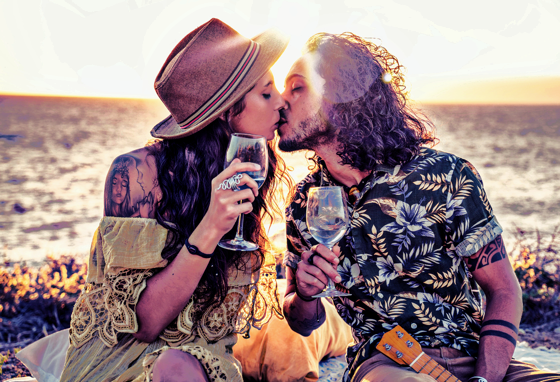 doi indragostiti se saruta pe plaja si au in mana pahare cu vin