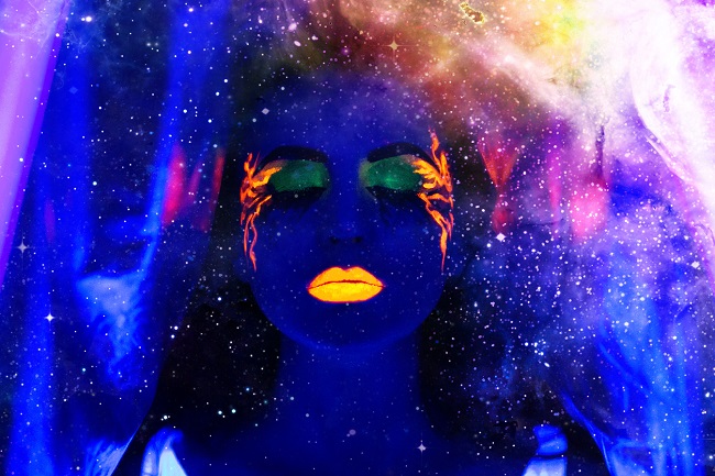 o femeie machiata cu culori neon sta cu ochii inchisi, pe fundal multe luminite