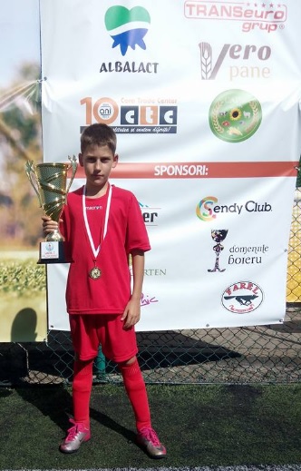 Fabian Căprău copil în echipament sportiv roșu ține o cupă în mână cu spatele la un banner sportiv
