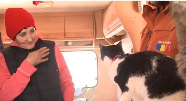 Doamna Ioana, femeia care are grijă de 50 de pisici, îmbrăcată cu hanorac roz, vestă neagră de fâș, având fes roșu pe cap,uitându-se la două dintre pisicile ei și ținându-și o mână pe piept