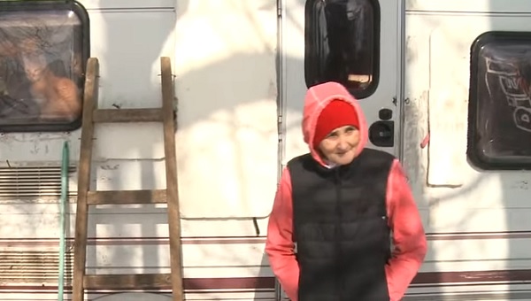 Doamna Ioana, femeia care are grijă de 50 de pisici într-o rulotă, îmbrăcată într-o bluză de trening roz cu glugă și cu o vestă de fâș neagră, stând în fața rulotei sale albe cu mâinile în buzunare