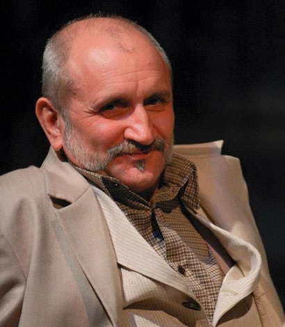 actorul Șerban Ionescu zambitor, imbracat intr-un costum bej, cu camasa cu patratele maro și bej